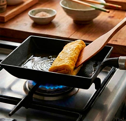 Omelette being cooked in a small black pan on a gas stove with a wooden spatula.